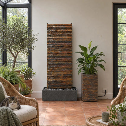 AVA GRANDE Wasserwand aus Schiefer Rusty in einem Wintergarten neben Korbsesseln und Pflanzen - CLIMAQUA (KI-Inhalte)