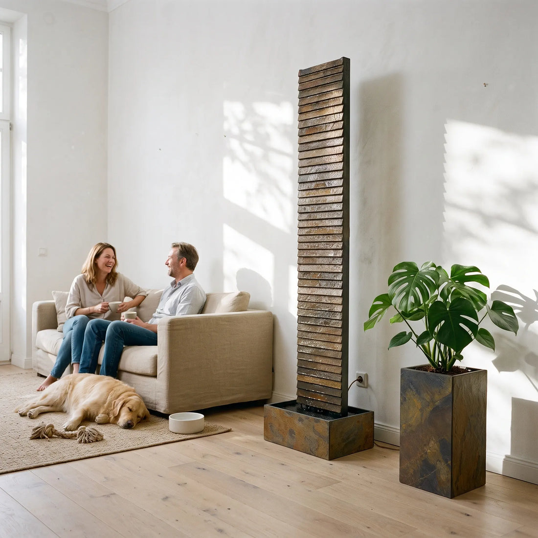 CLIMAQUA AVA XL Wasserwand aus Schiefer im Wohnzimmer neben einem Sofa, daneben ein PYLO 60 Pflanzgefäss mit Monstera