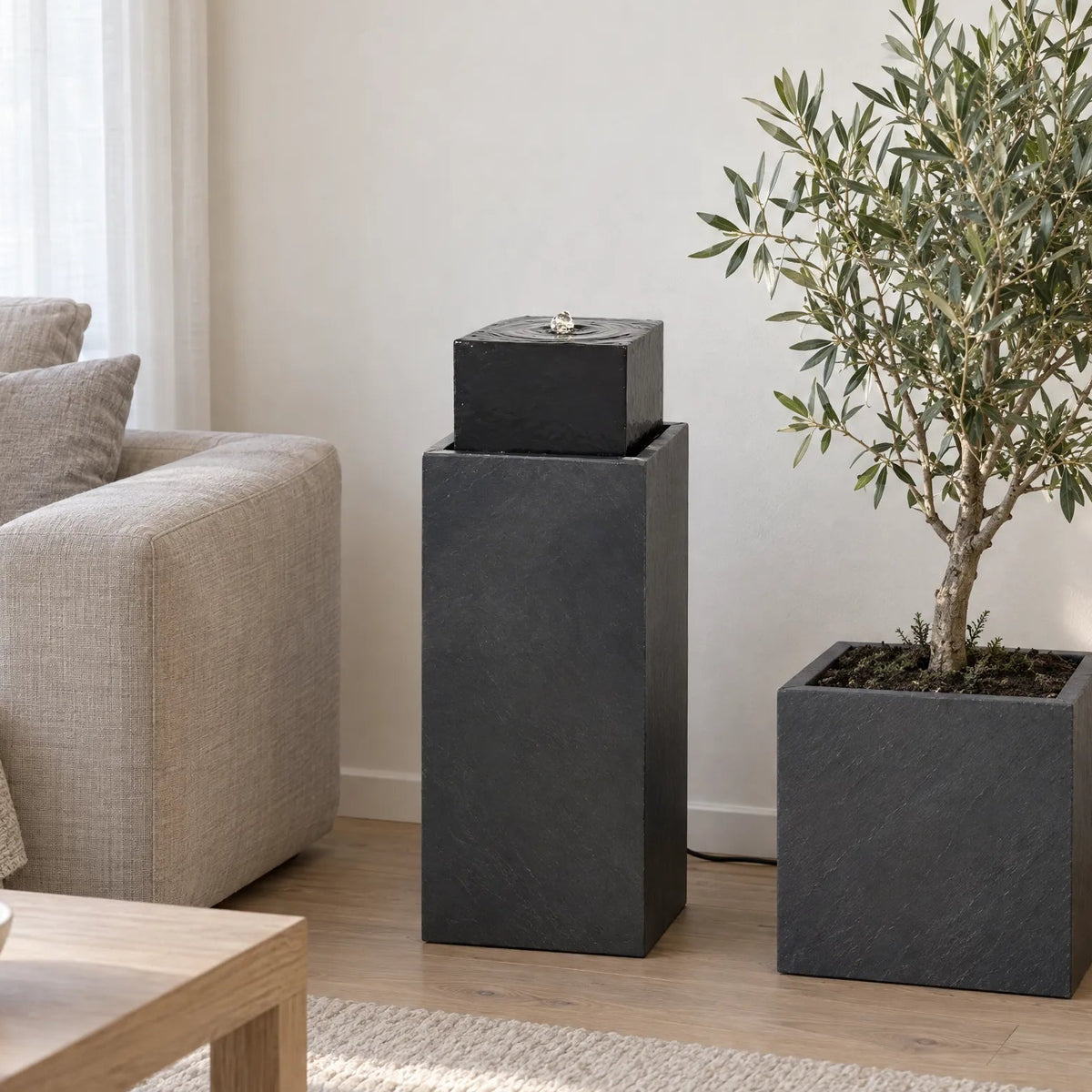 Säule Brunnen NEO Anthrazit im Wohnzimmer neben einem Sofa, daneben ein Pflanzgefäss mit Olivenbaum - CLIMAQUA